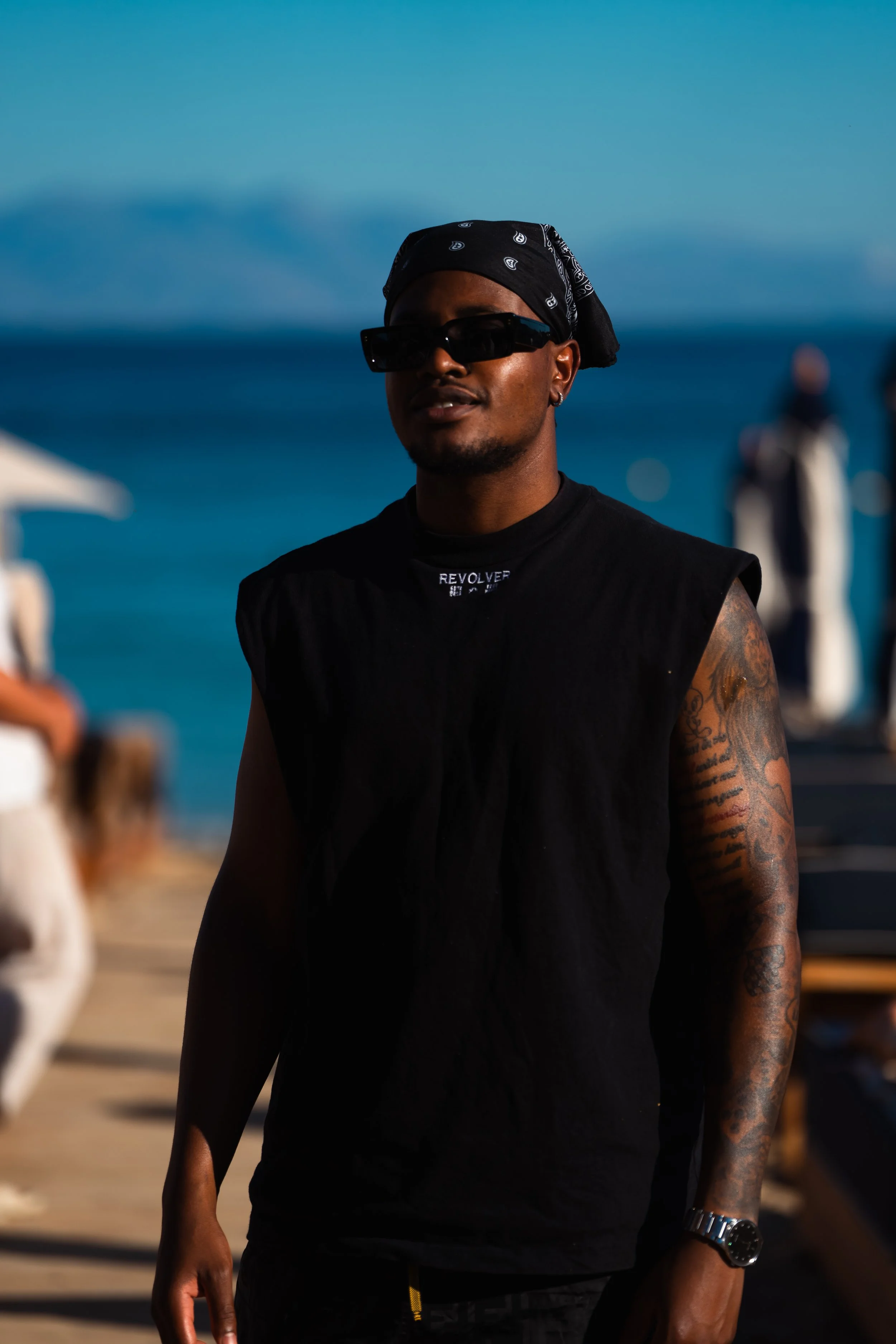 A man with dark skin, wearing sunglasses, a black sleeveless shirt, a black bandana, and a wristwatch, stands on a beach with the ocean and mountains in the background.