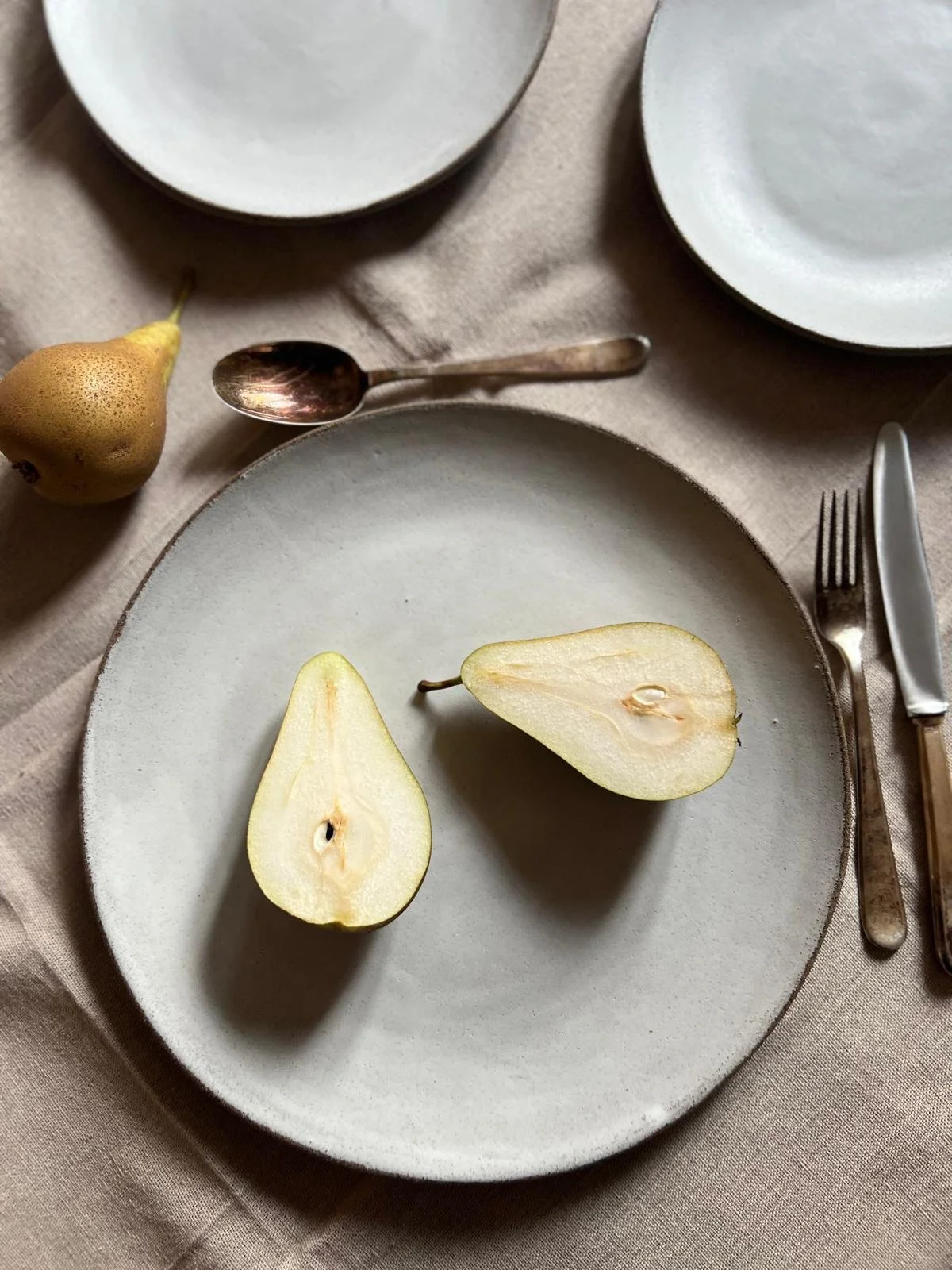 A beige tablecloth with a ceramic plate holding a halved pear, with other empty plates, a spoon, a fork, and a knife on the table.