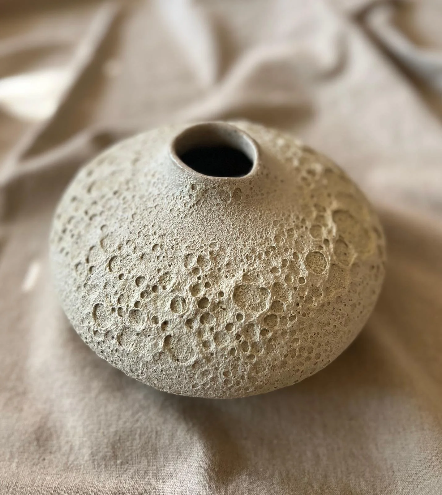 A round ceramic vase, porous stone resembling a small volcano or the moon texture, placed on a soft, light-colored fabric surface.
