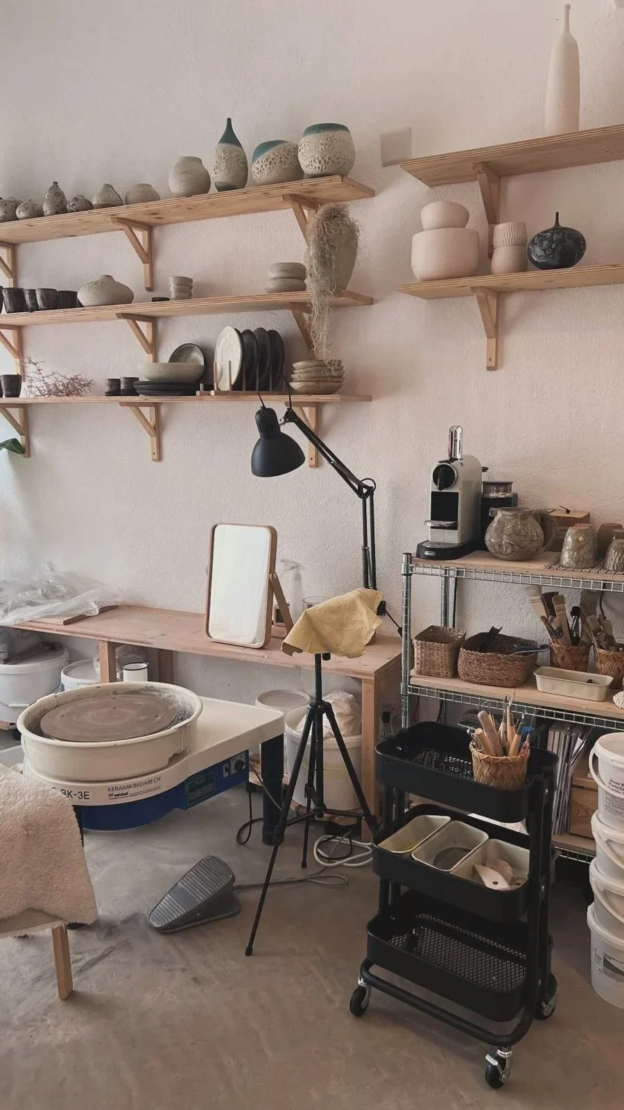 A pottery studio with wooden shelves holding various pottery pieces, a work table with pottery equipment, and a black rolling cart filled with brushes and tools.