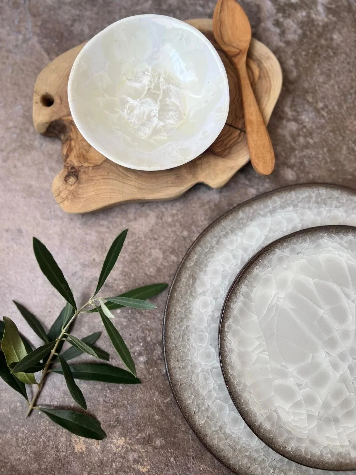 Two round white plates with a textured pattern, a branch of green leaves, a small bowl with a white substance, and a wooden spoon on a textured surface.