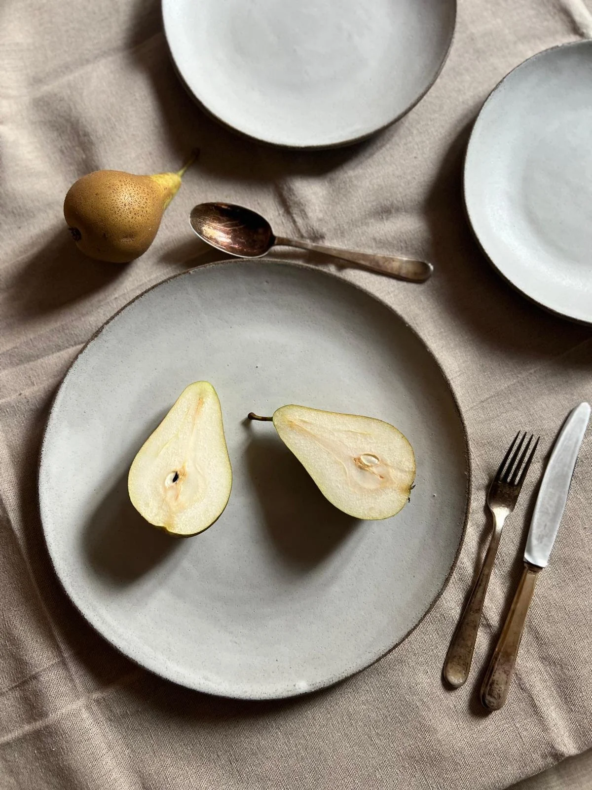 A table setting with two halves of a pear on a gray ceramic plate, a whole pear, a spoon, a fork, and a knife on a beige tablecloth.
