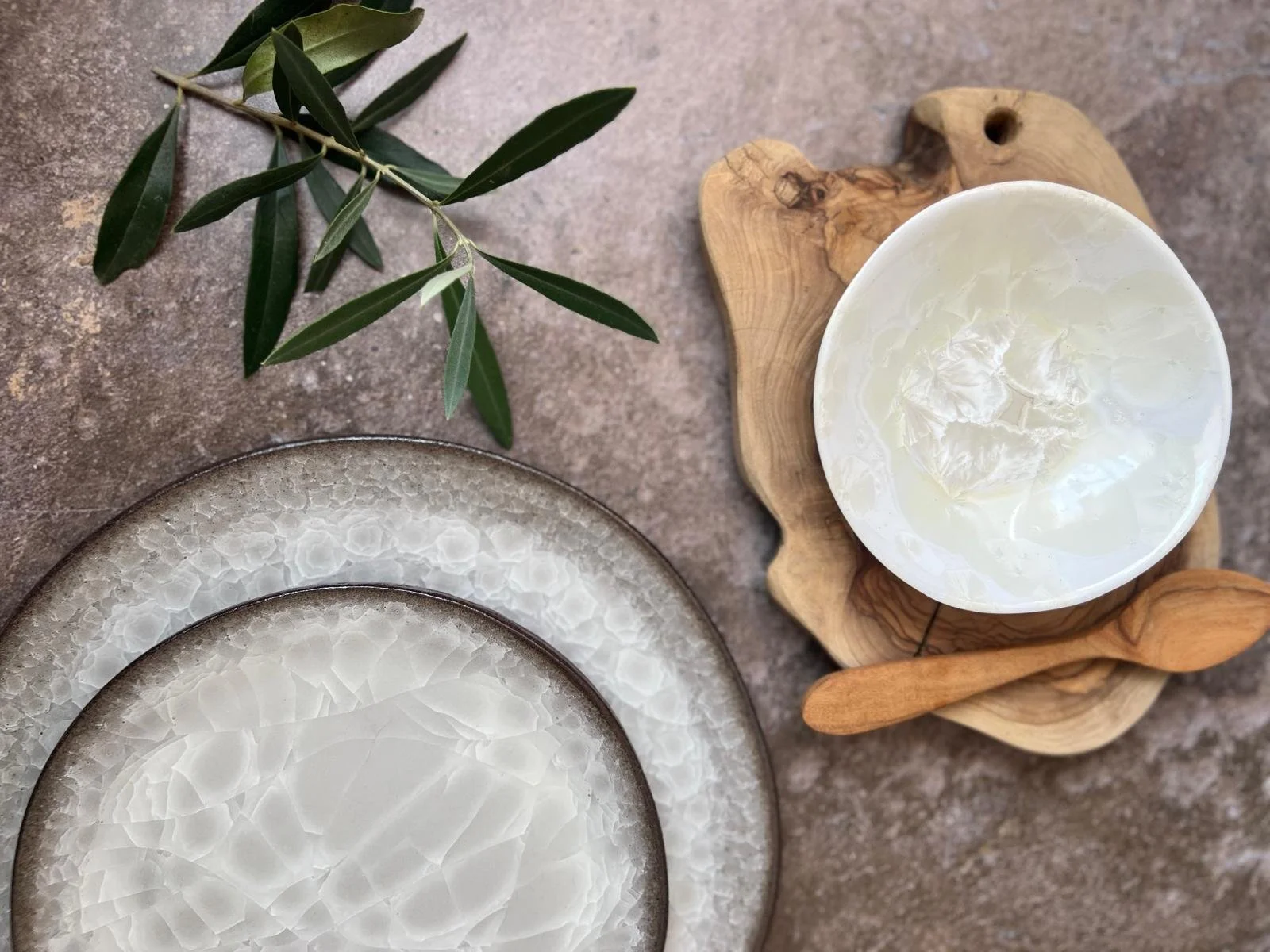 White ceramic bowl on a wooden tray, with two ceramic plates with textured patterns, and a branch of green leaves on a brown textured surface.