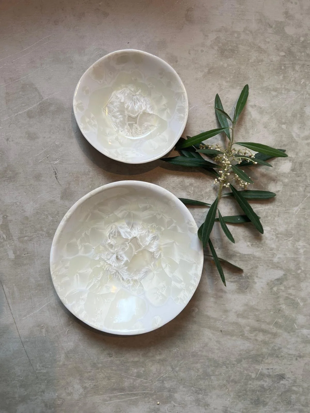 Two white ceramic bowls with a marbled pattern on a gray surface, accompanied by green leaves and small white flowers next to the bowls.