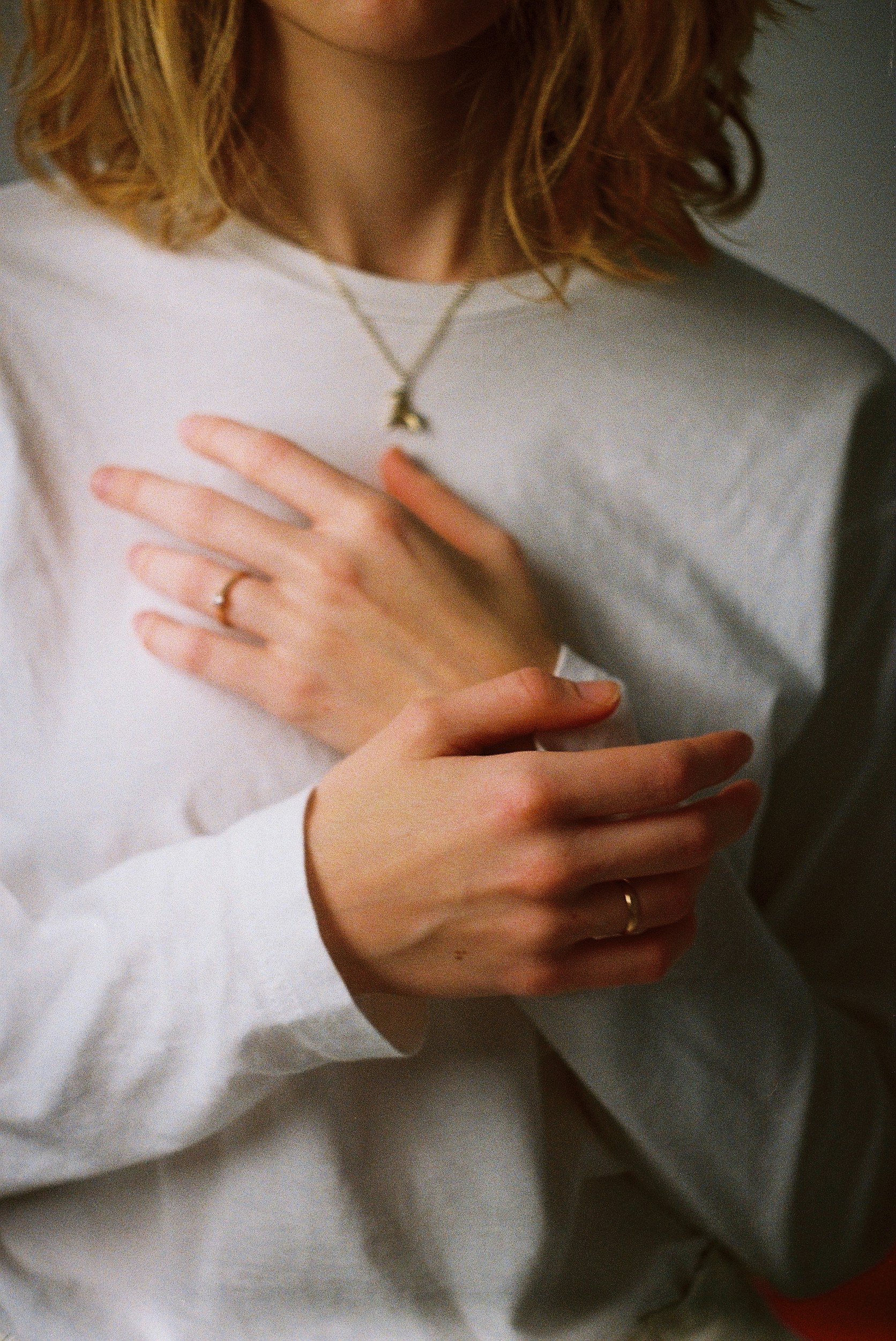 Close-up of a woman wearing a white shirt, with her hands crossed over her chest, displaying rings and a necklace.