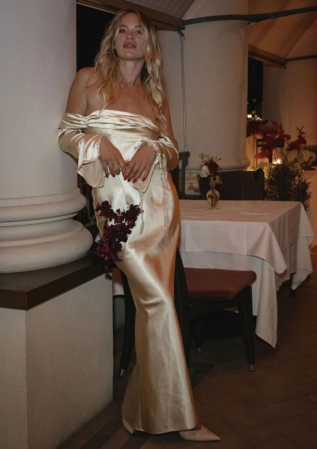 Eine Frau steht in einem weissen Kleid an einer Säule und hält eine Tasche aus Blumen.