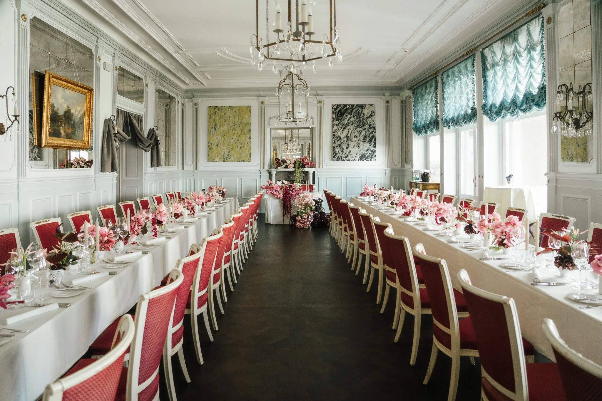 In einem Raum stehen zwei lange weisse eingedeckte Tische mit roten Stühlen und einer Blumendekoration in pink.
