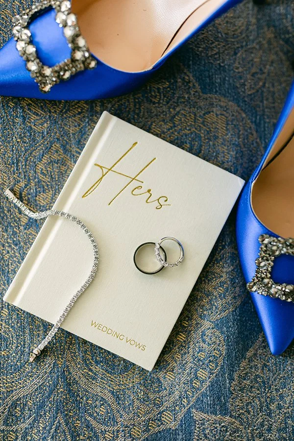 Auf einem beigen Buch liegen zwei Ringe und ein Armband und daneben stehen zwei blaue Highheels.