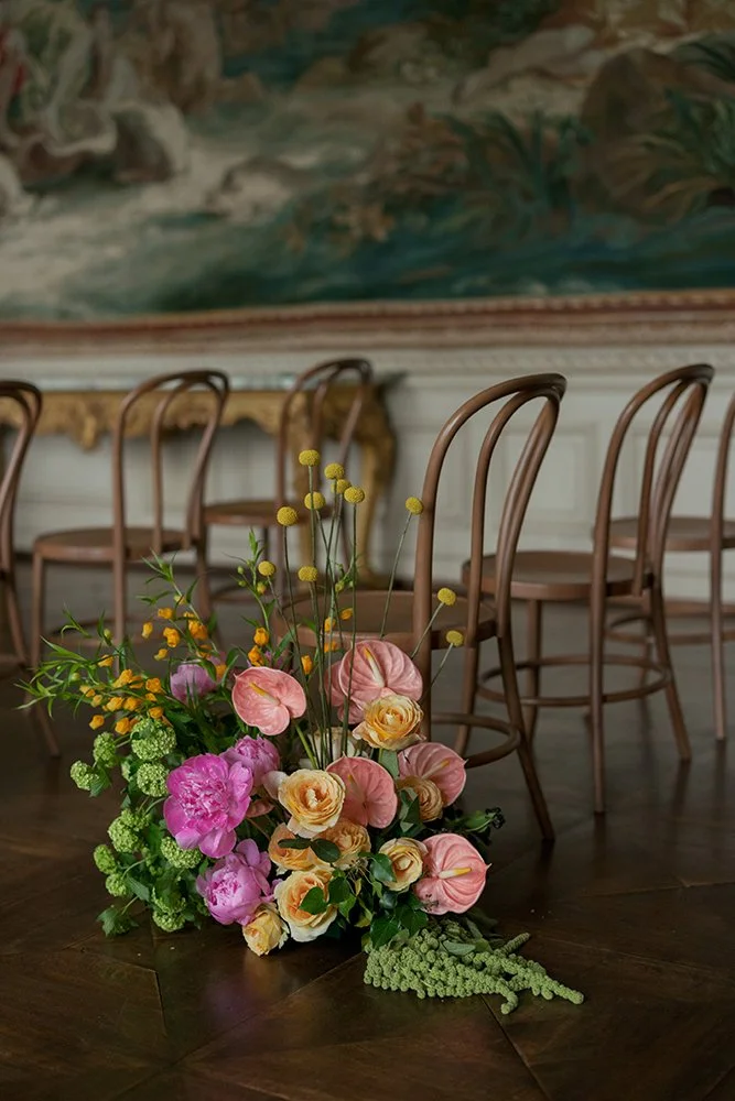 Ein Blumenarrangement steht am Boden vor Holzstühlen.