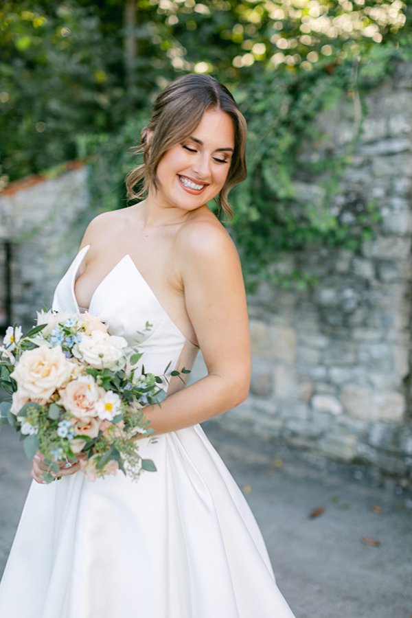 Eine Frau steht lachend in einem Garten in ihrem Brautkleid mit ihrem Brautstrauss.