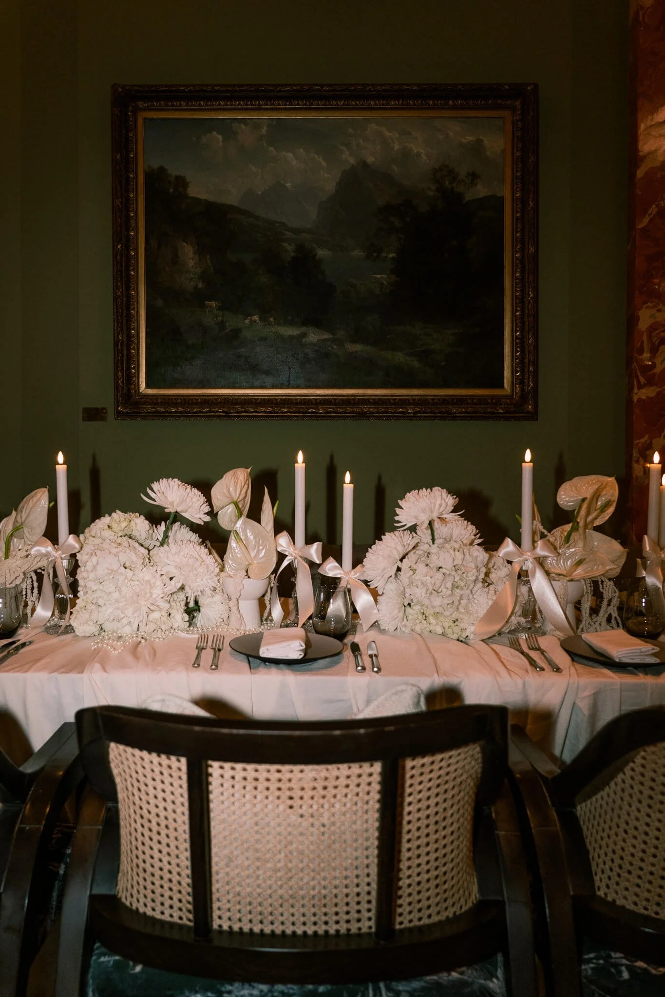 Ein Tisch ist dekoriert mit weissen Blumen und Stabkerzen vor einer Wand mit Bild.