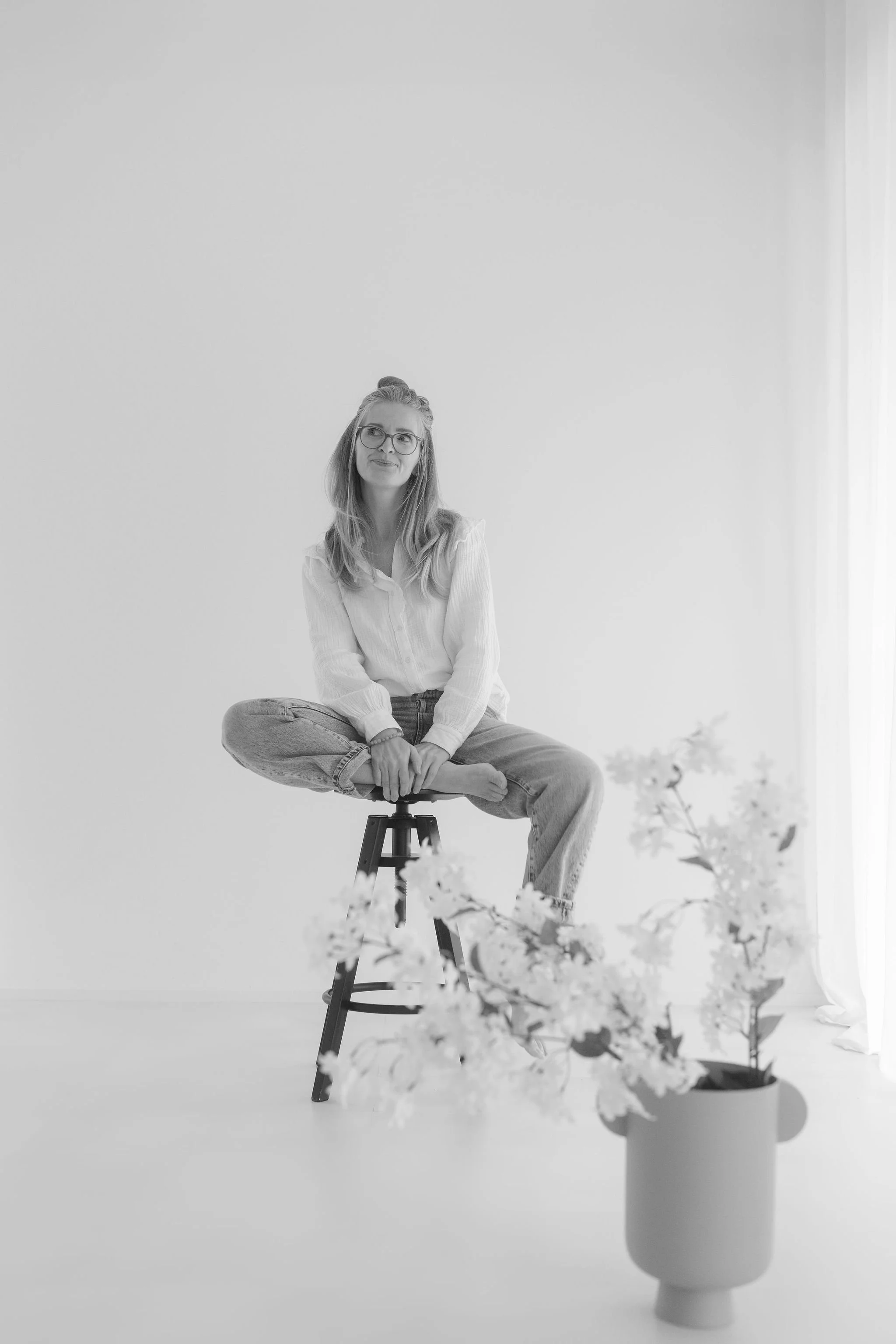 Schwarz-Weiß-Foto einer jungen Frau mit Brille, sitzend im Schneidersitz auf einem Barhocker vor einer weißen Wand, mit Blumen im Vordergrund.