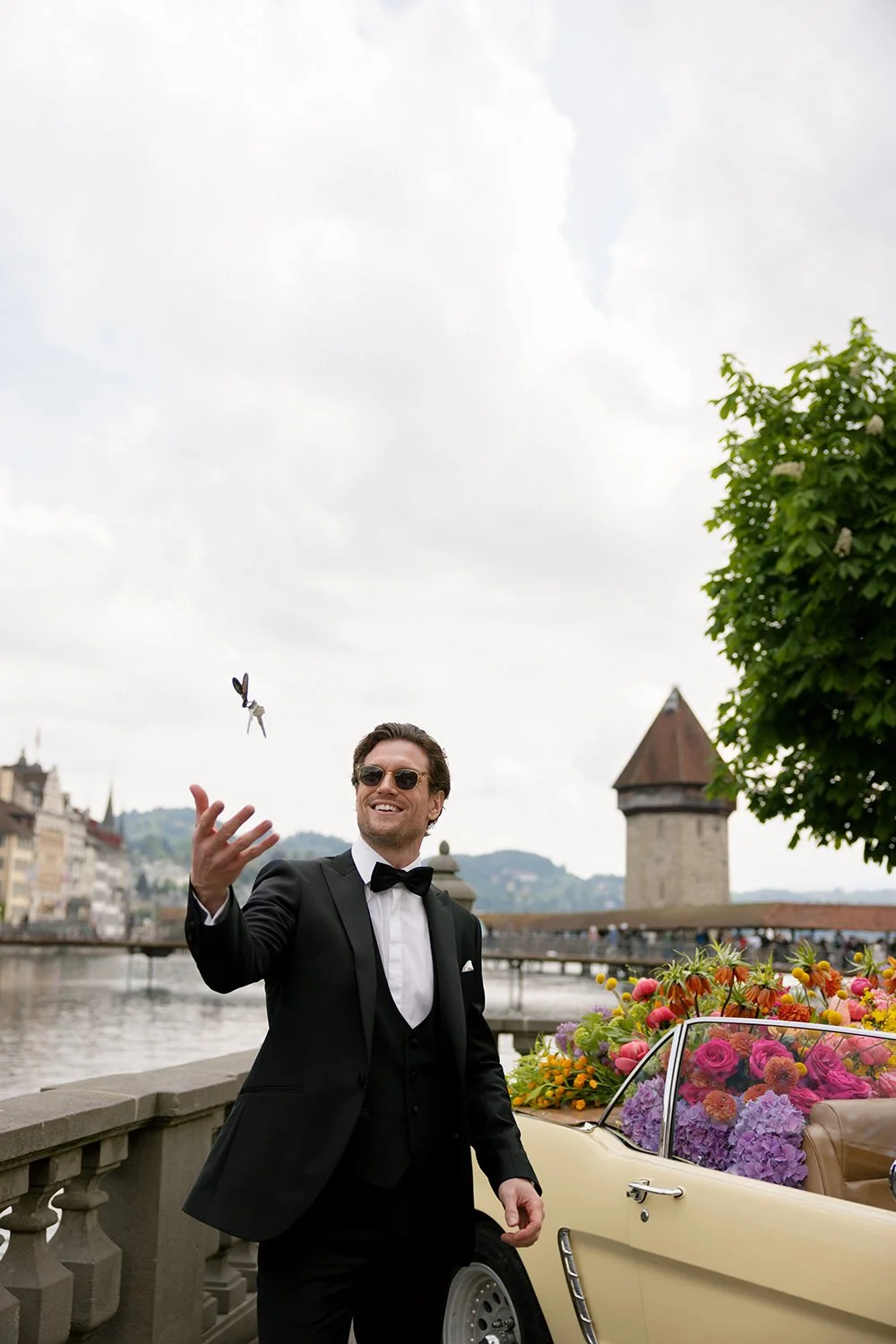 Ein Mann im schwarzen Anzug steht neben einem alten Auto und wirft einen Schlüssel in die Luft.