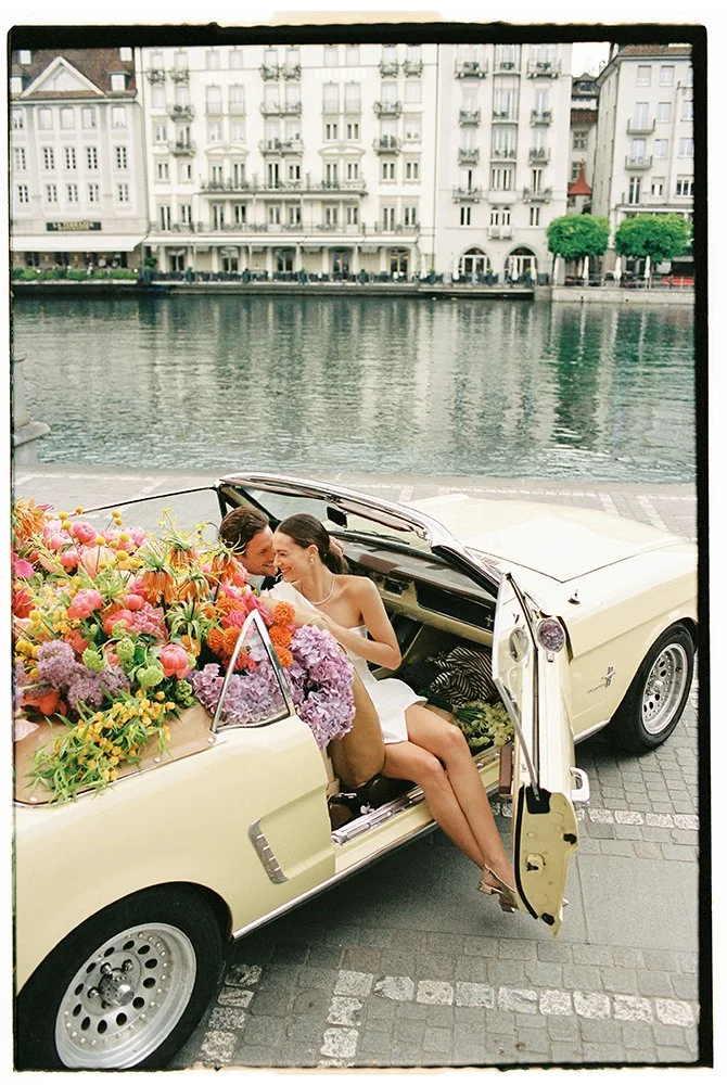 Ein Brautpaar sitzt in einem hellgelben Oldtimer, der gefüllt ist mit bunten Blumen.