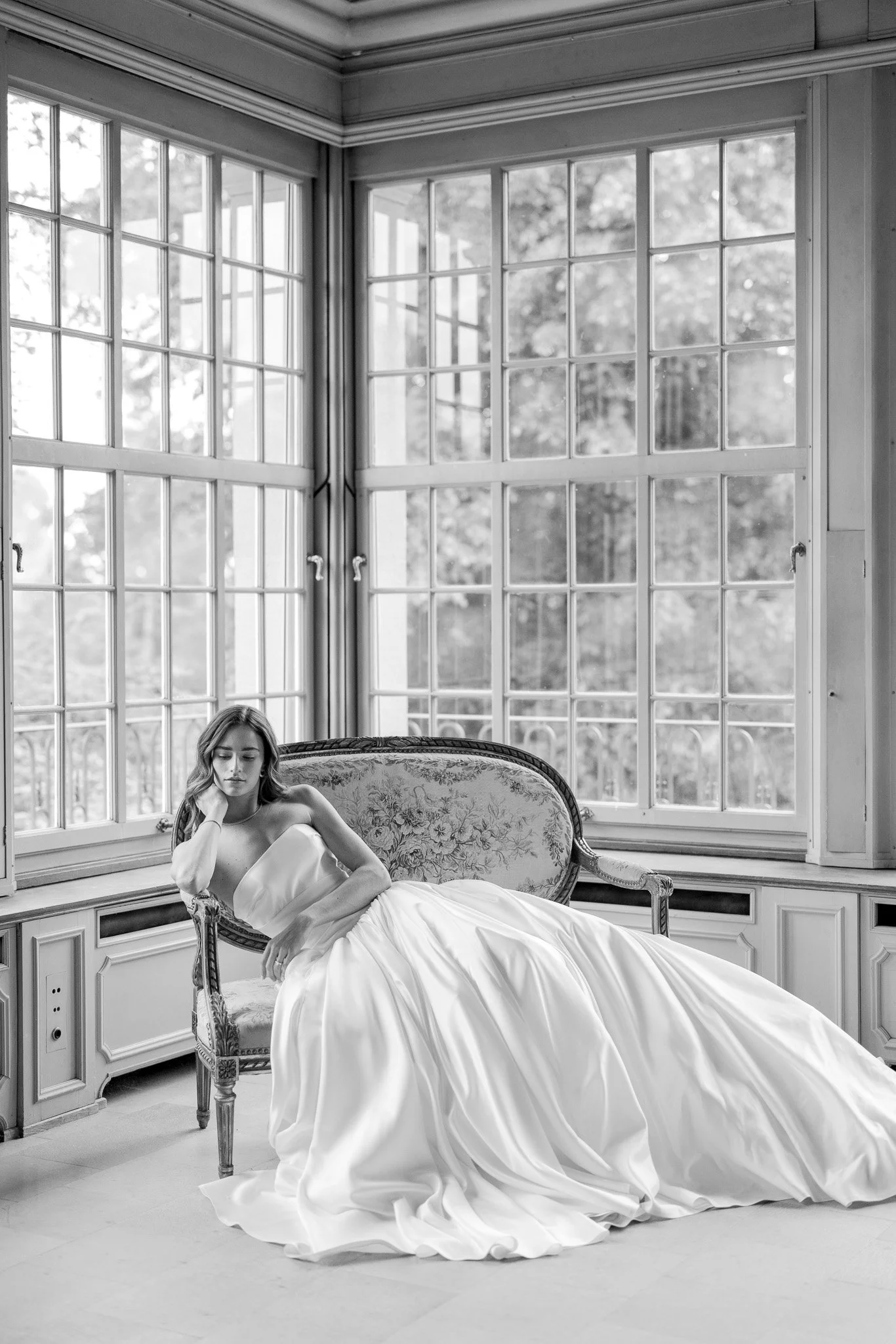 Eine junge Frau in einem lang weißen Brautkleid liegt auf einem alten, eleganten Sofa in einem Raum mit großen Fensterfronten. Schwarz-Weiß-Fotografie.