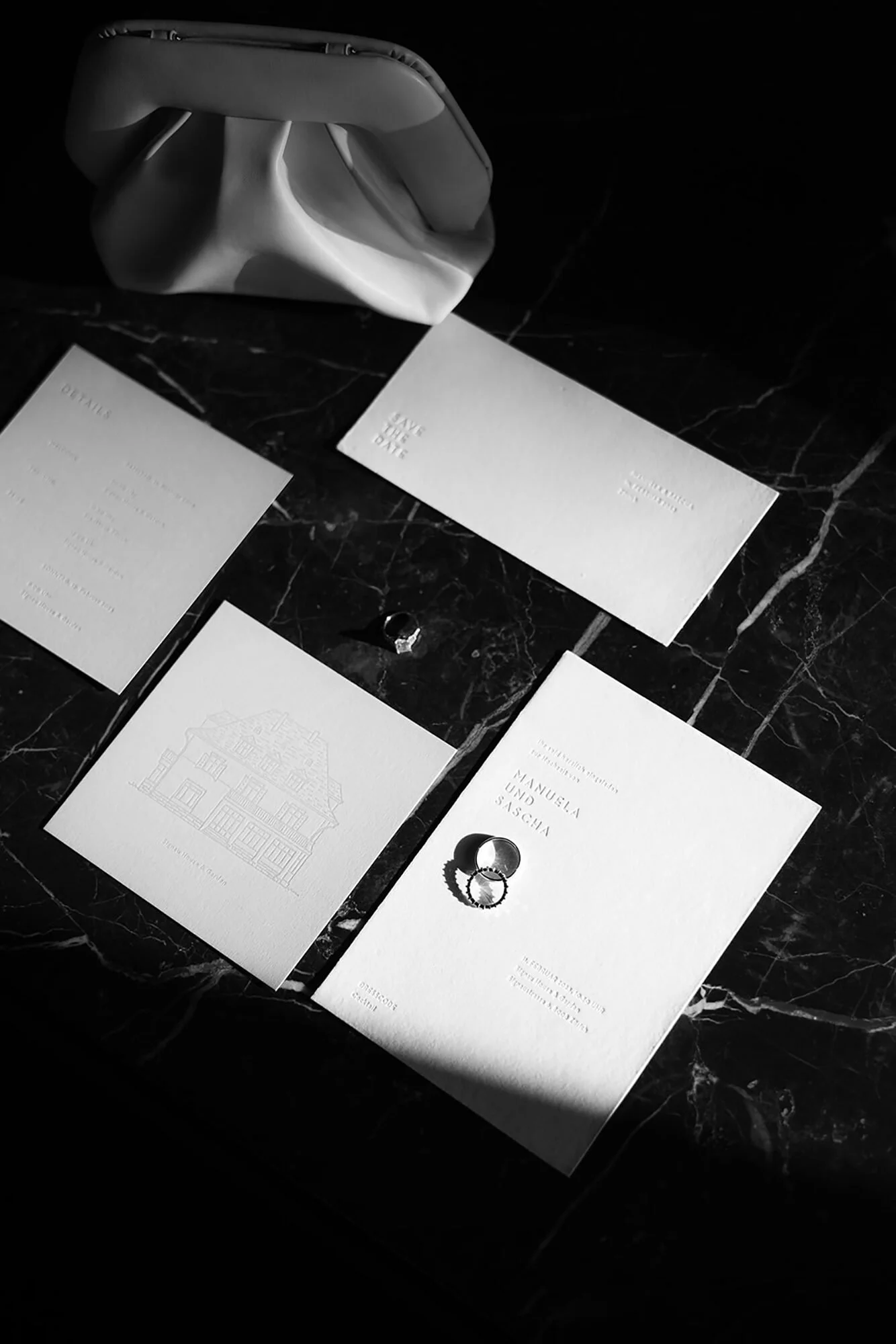 Schwarz-weiße Hochzeits-Einladungskarten, zwei Ringe, eine weiße Tasche auf dunklem Marmor-Tisch.