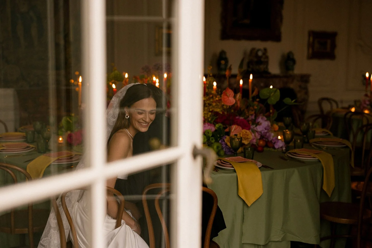 Hinter einem Fenster sitzt eine lachende Braut vor einem Tisch mit Blumen.