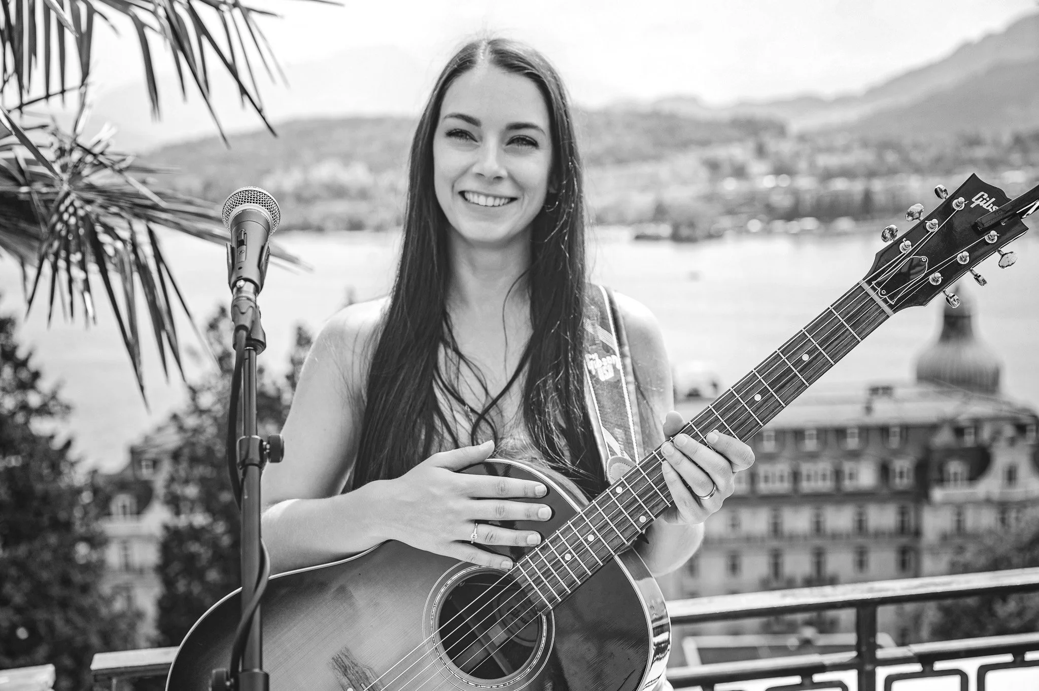Eine junge Frau mit langen Haaren spielt auf einer Gitarre und singt in einem Outdoor-Umfeld, mit Wasser und Bergen im Hintergrund. Sie hat ein Lächeln auf dem Gesicht und steht vor einem Mikrofon.