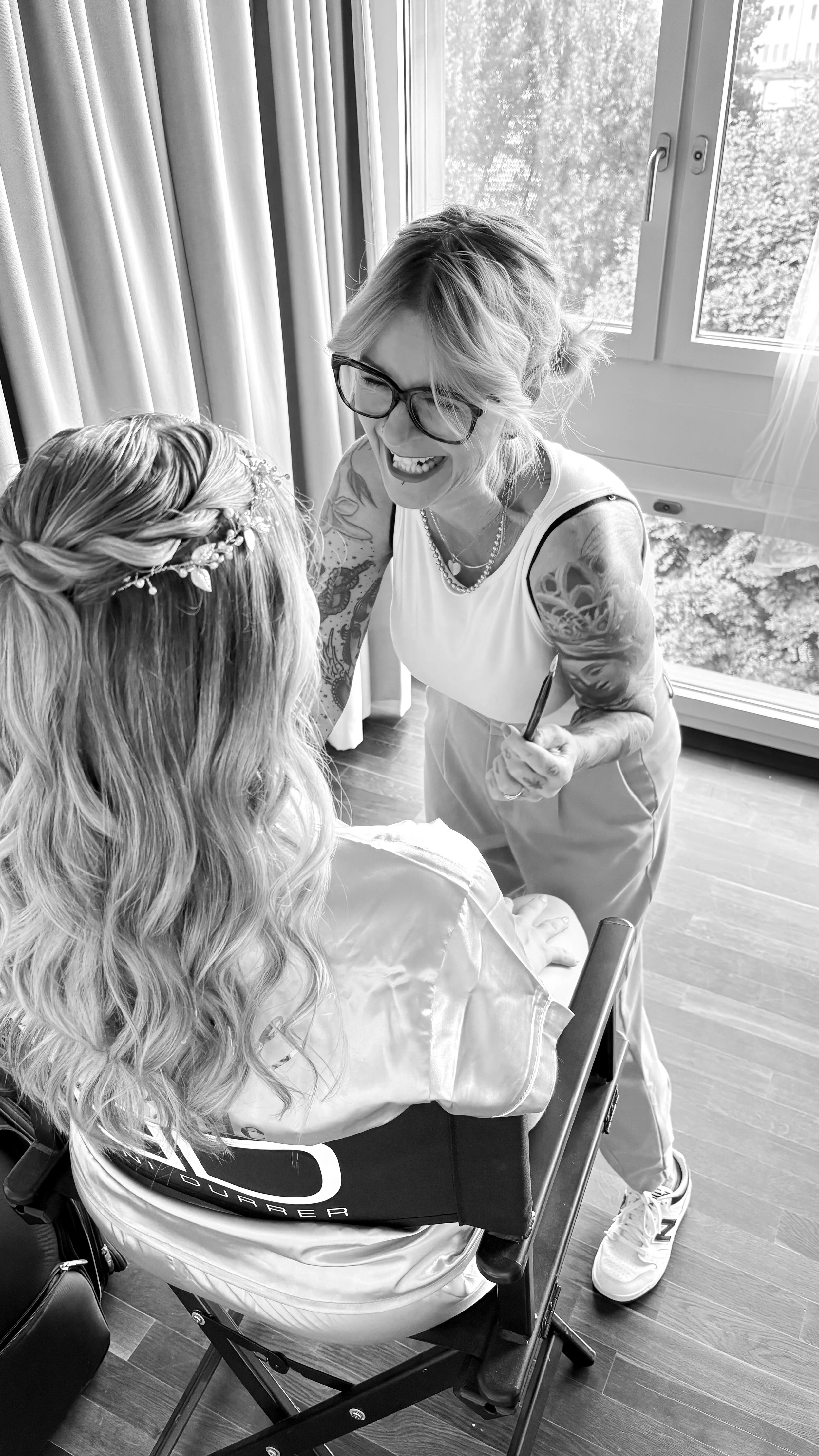 Eine lachende Frau mit Kurzhaarschnitt und Tattoos, spricht mit einer sitzenden Frau, die eine dekorative Haarpracht trägt, in einem Raum mit großen Fenster und Vorhängen.