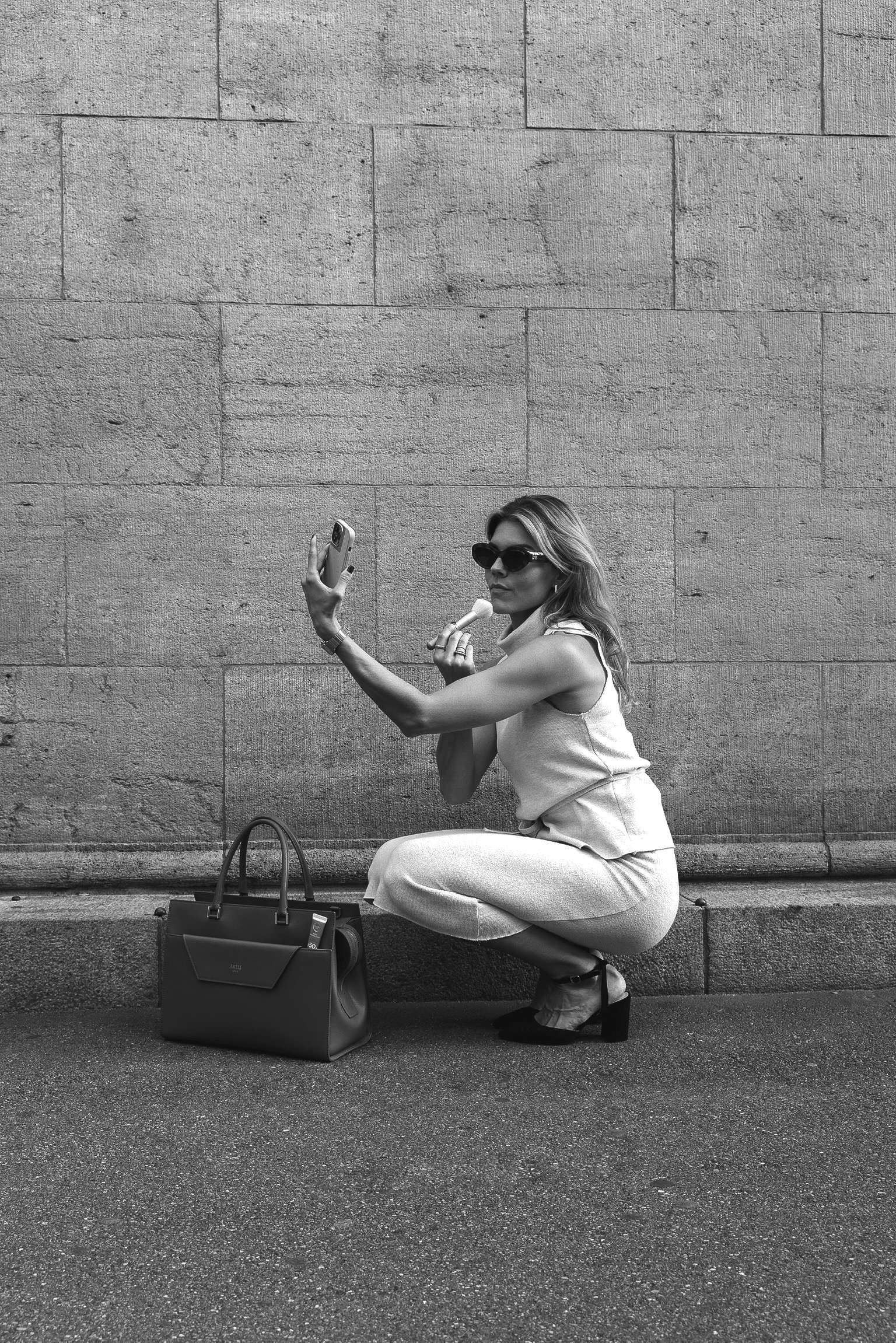 Frau in Sonnenbrille, sitzend an einer Steinmauer, fotografiert sich mit Smartphone, hält Eiskugel, neben Handtasche auf dem Boden.