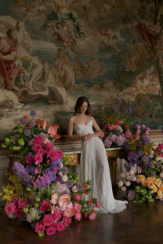 Eine Frau im weissen Kleid steht vor einer Wand umgeben von bunten Blumen.