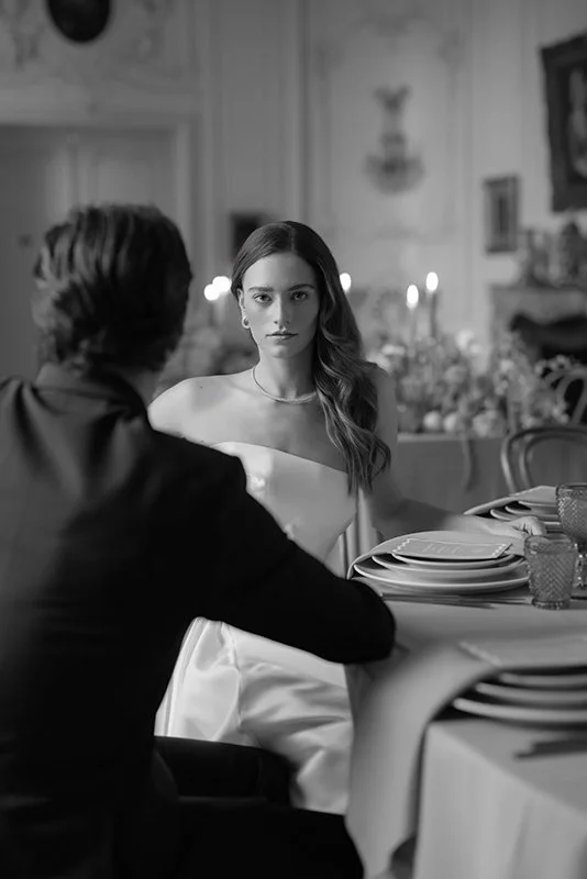 Eine Frau sitzt hinter einem Mann an einem Tisch und schaut ernst in die Kamera.