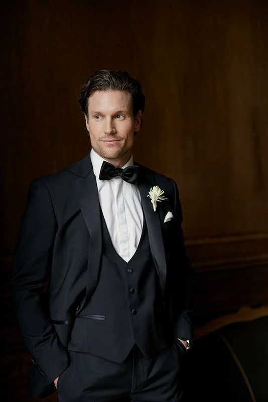 Ein Mann steht in einem schwarzen Anzug vor einer Holzwand.