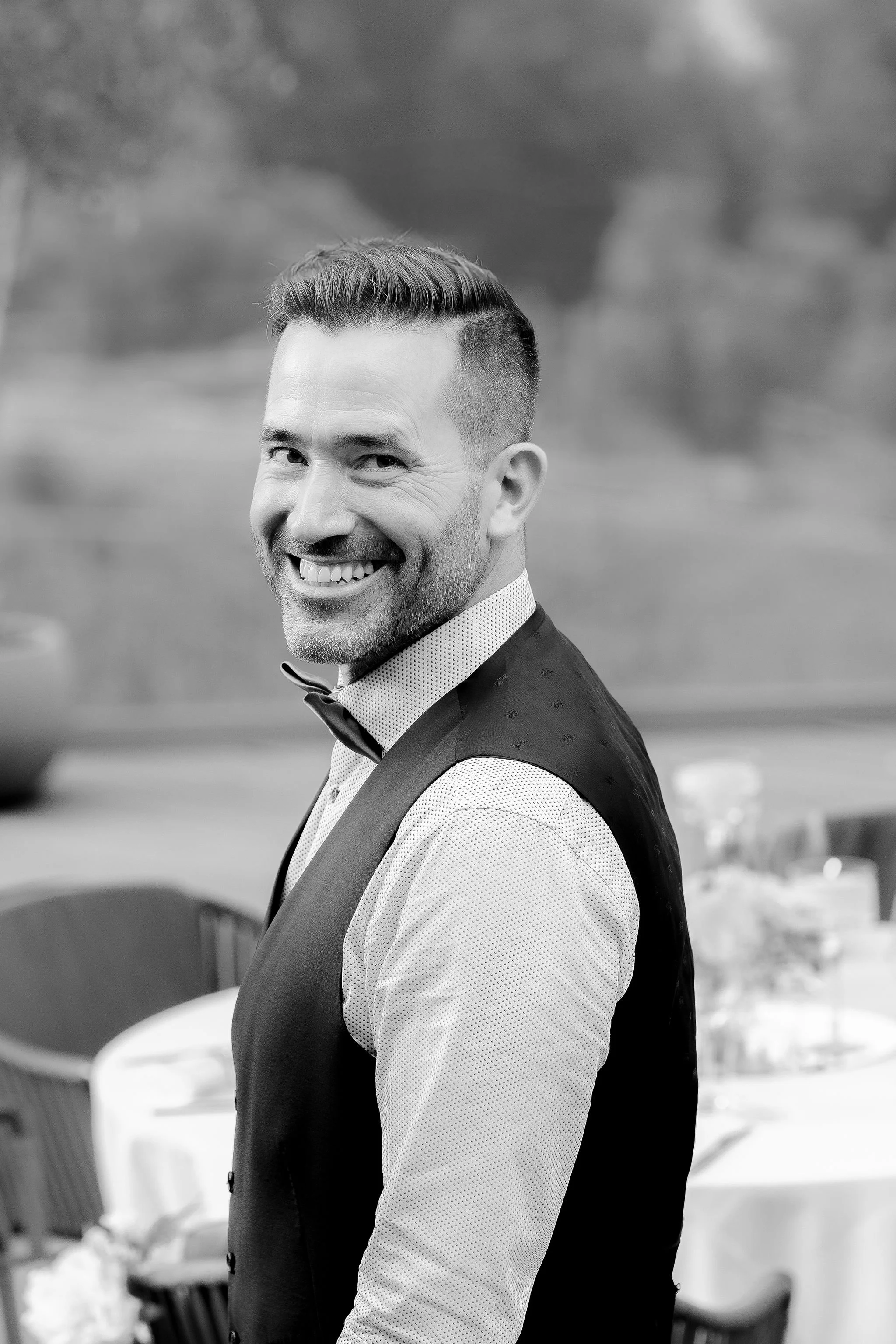 Lächelnder Mann in formeller Kleidung bei einer Veranstaltung, im Hintergrund gedeckter Tisch, schwarz-weiß.