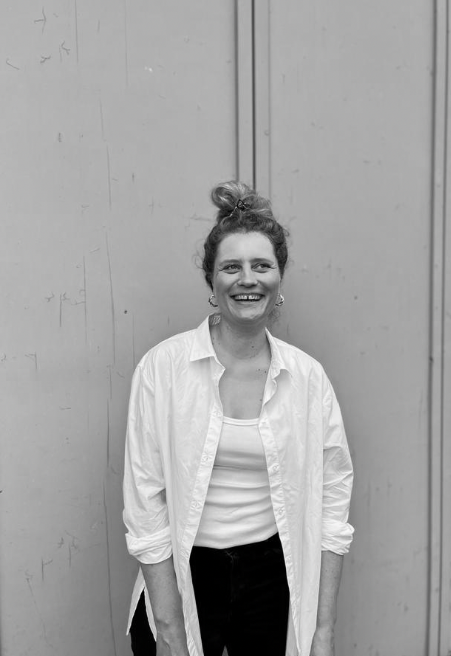 Eine lachende Frau mit lockigem Haar in einem hohen Dutt, trägt eine weiße Bluse, steht vor einer grauen Wand und lächelt in die Kamera.