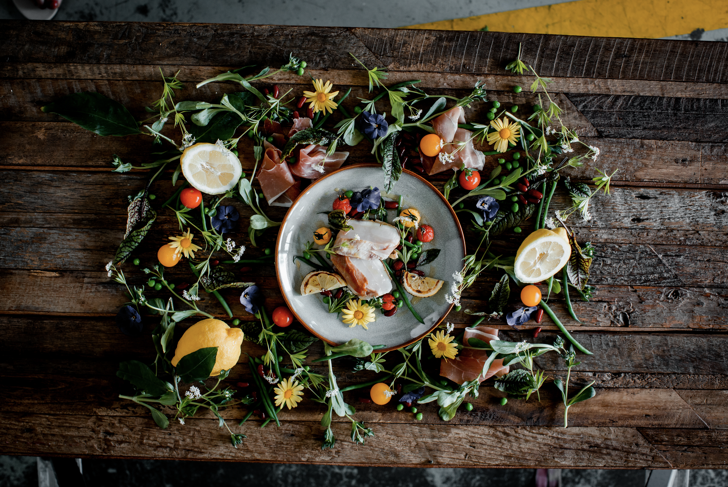 Boden aus Holz mit einer roten und weißen Speiseplatte in der Mitte, umgeben von Zitronenscheiben, bunten Erbsen, Blättern, Blumen und Schinken, alles dekorativ arrangiert.