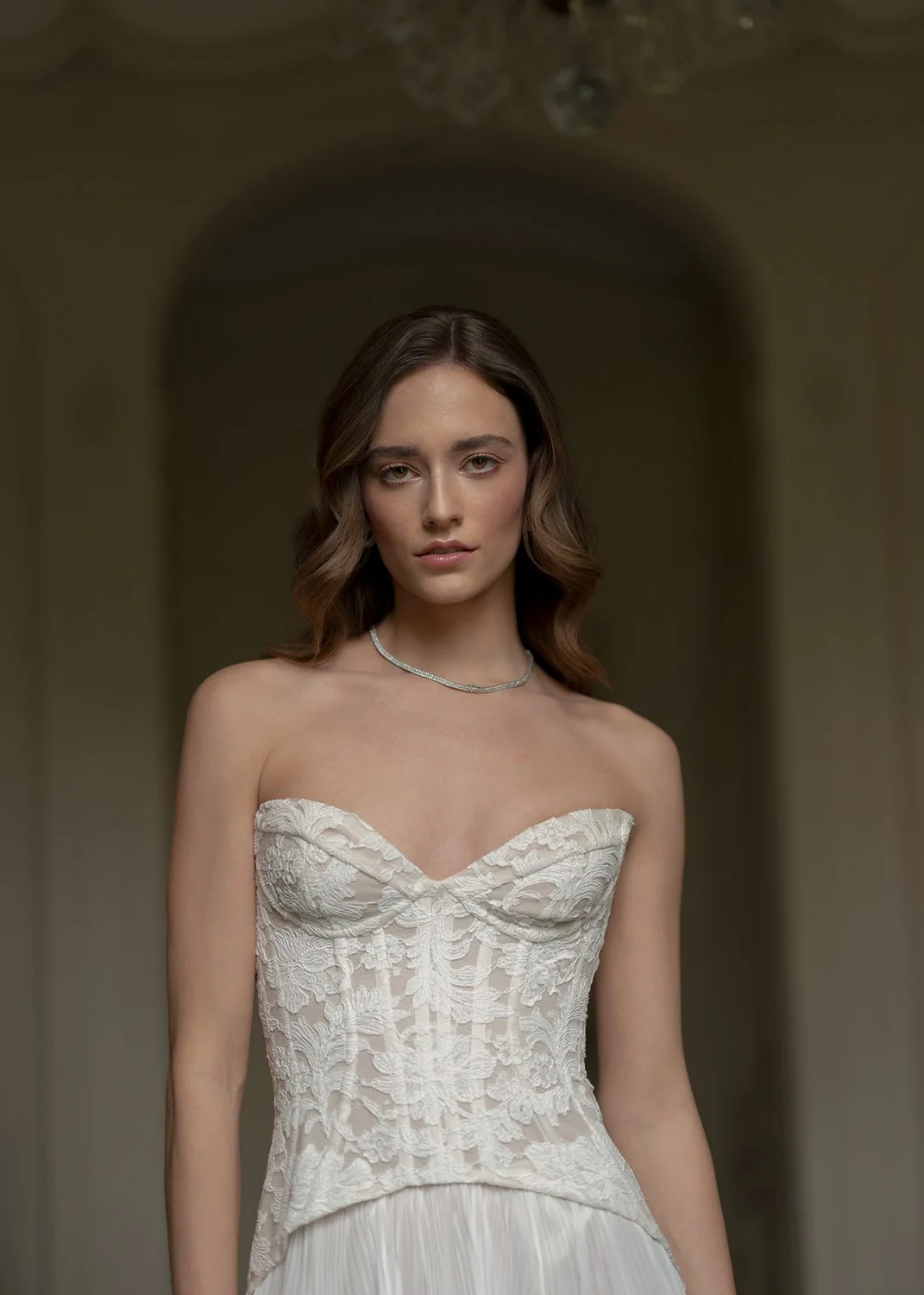 Eine Frau steht in einem weissen Kleid in einem Raum und schaut selbstbewusst in die Kamera.