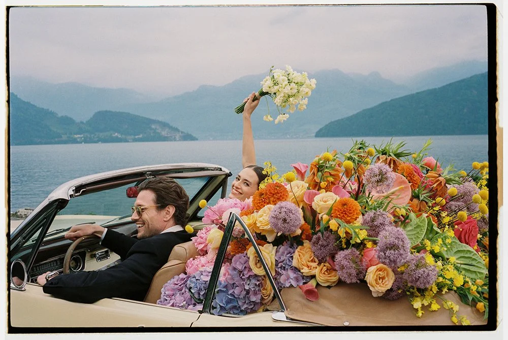 Ein Paar sitzt in einem Oldtimer gefüllt mit bunten Blumen am Vierwaldstättersee.