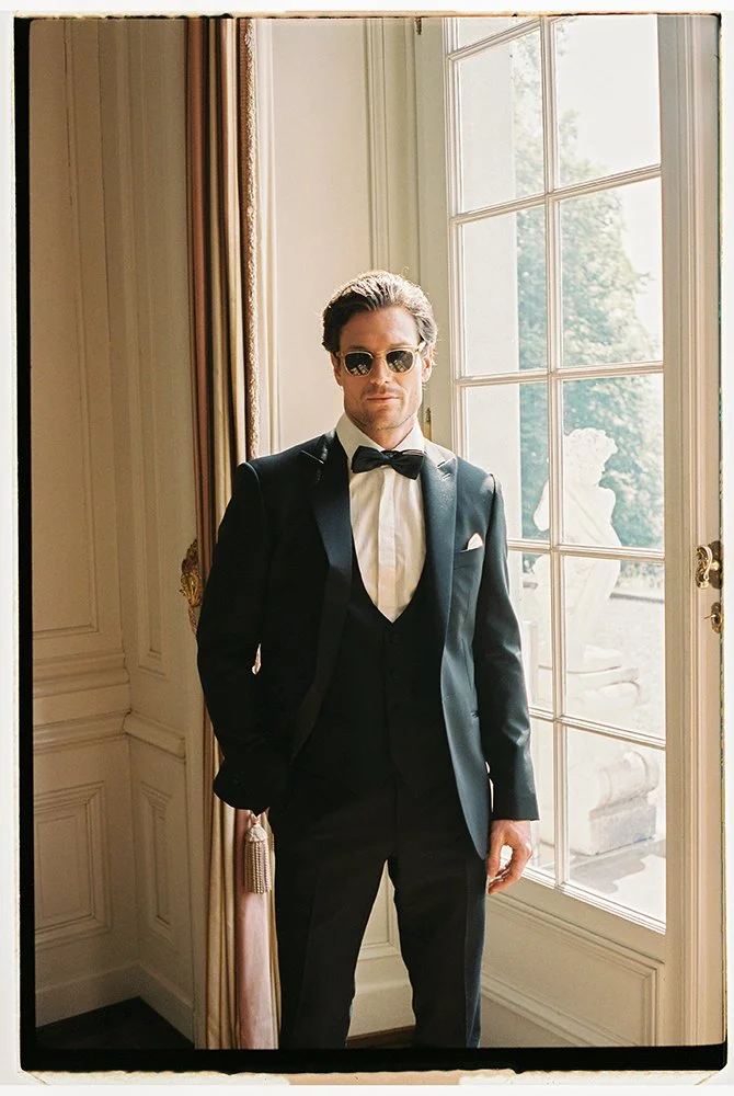 Ein Mann steht in einem schwarzen Anzug und Sonnenbrille vor einem Fenster.