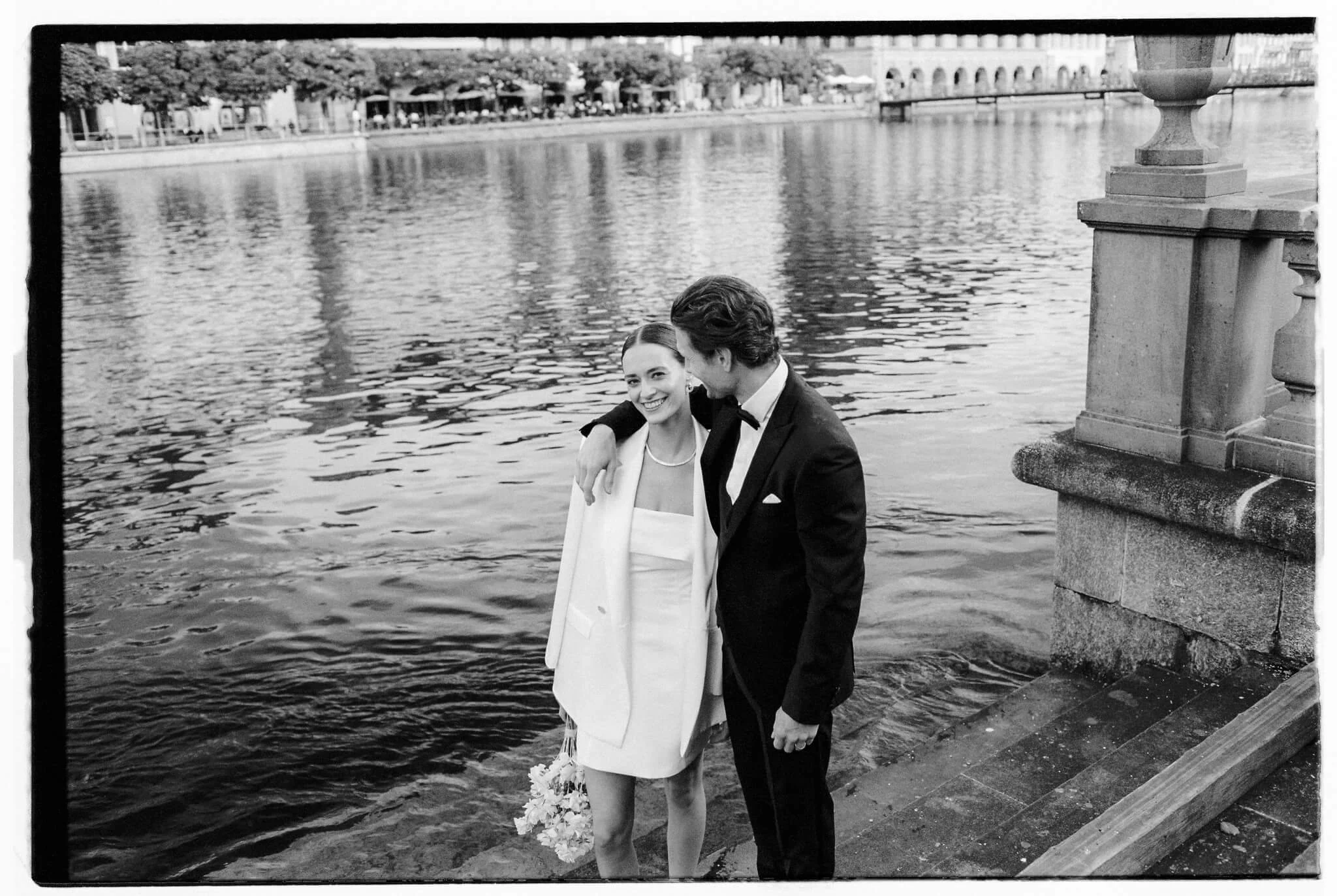 Ein Brautpaar steht lächelnd am Wasser mit einem Brautstrauss.