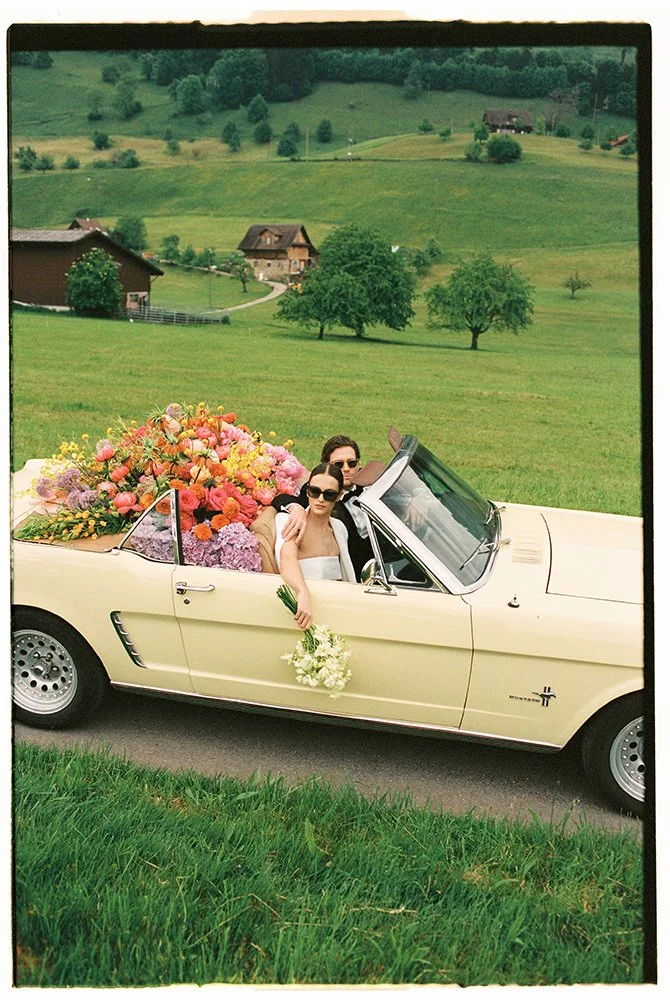 Ein hellgelber Oldtimer steht gefüllt mit bunten Blumen auf einer Wiese.