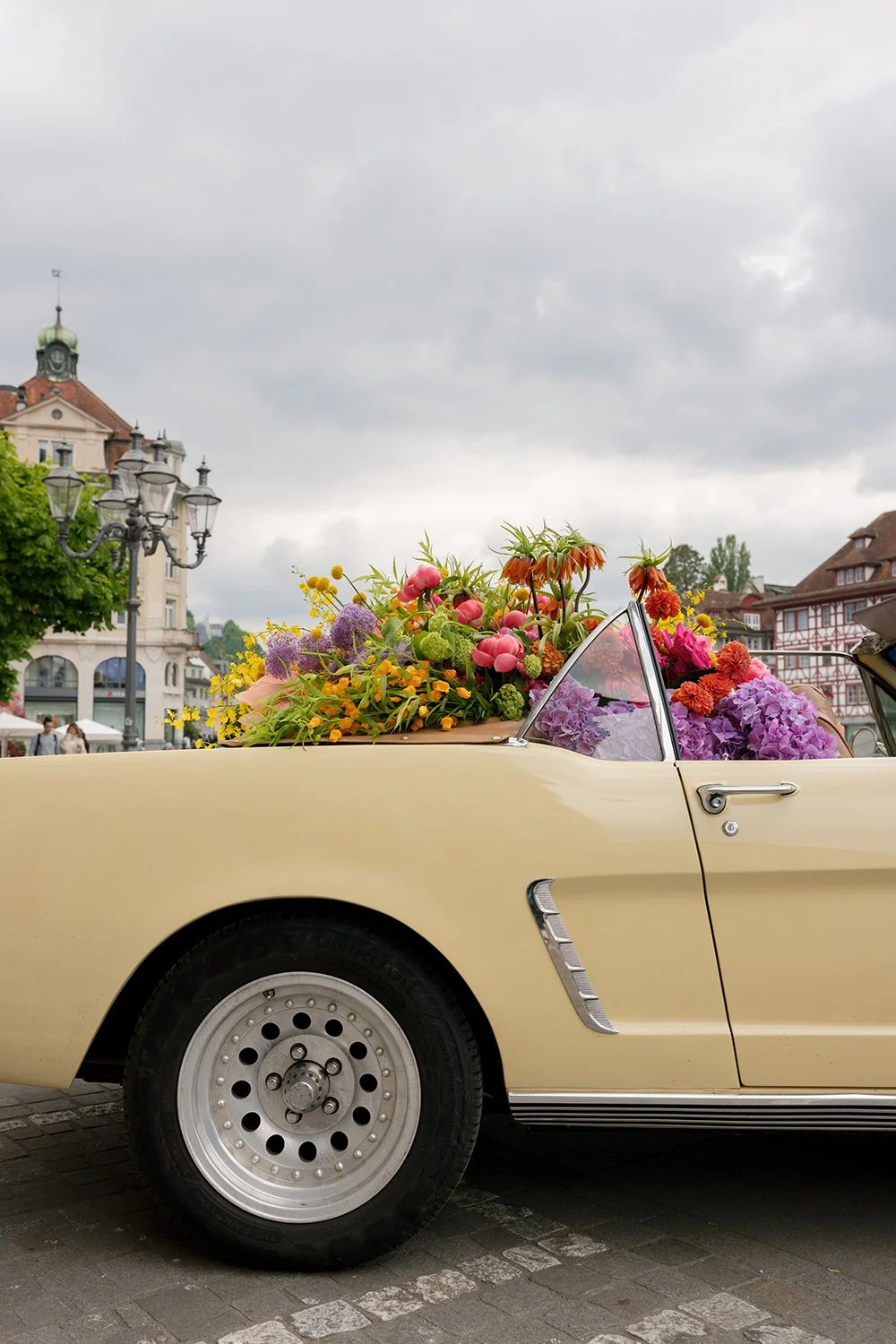 Ein hellgelber Oldtimer steht auf einem Platz und ist gefüllt mit bunten Blumen.