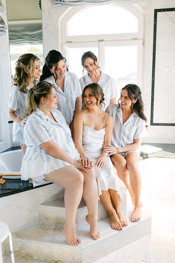 Sechs Frauen sitzen lachend nebeneinander in hellblauen Kleidern.