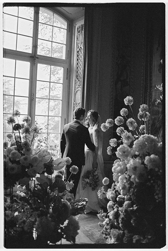 Ein Brautpaar steht vor dem Fenster umgeben von vielen Blumen.