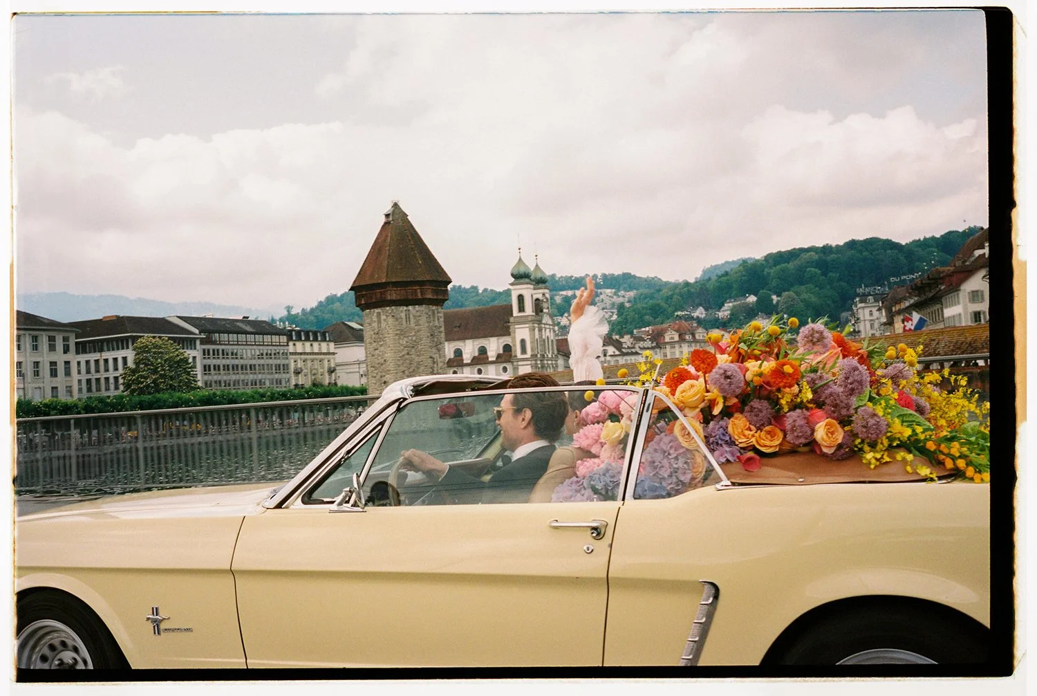 Ein hellgelber Oldtimer ist gefüllt mit bunten Blumen und fährt vor der Kappelbrücke.