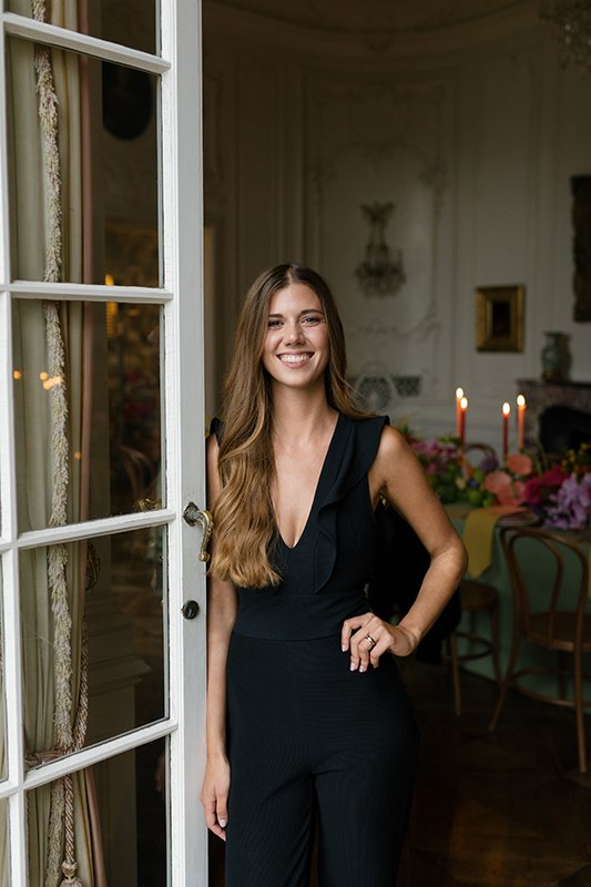 Eine Frau steht lackend in einem schwarzen Overall neben einem alten Fenster.