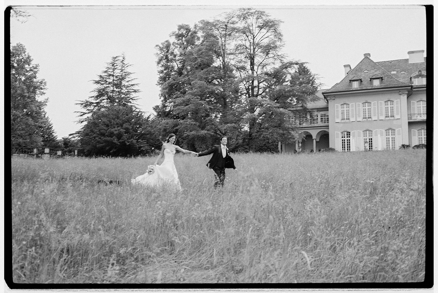 Ein Brautpaar springt verliebt auf einer Wiese vor einem alten Haus.
