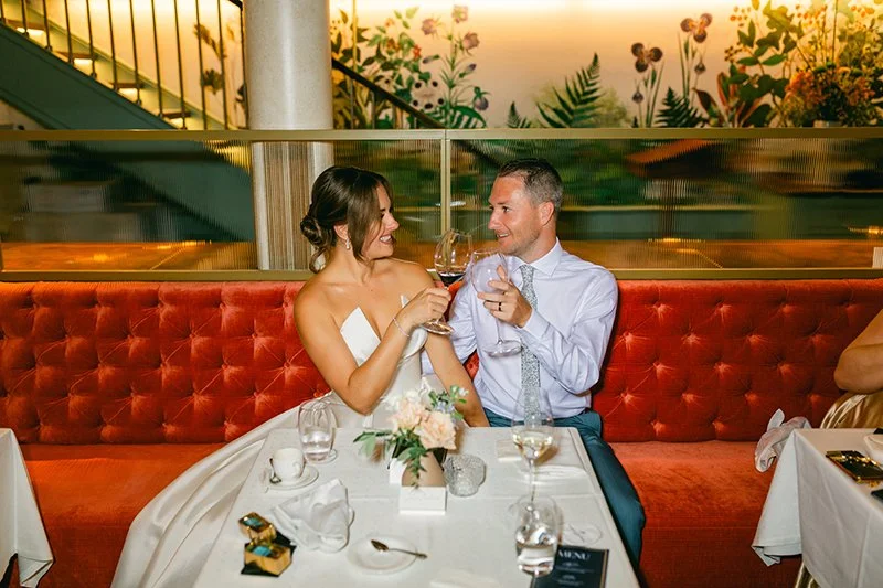 Ein Brautpaar sitzt auf einer roten Bank an einem Tisch und stosst an.