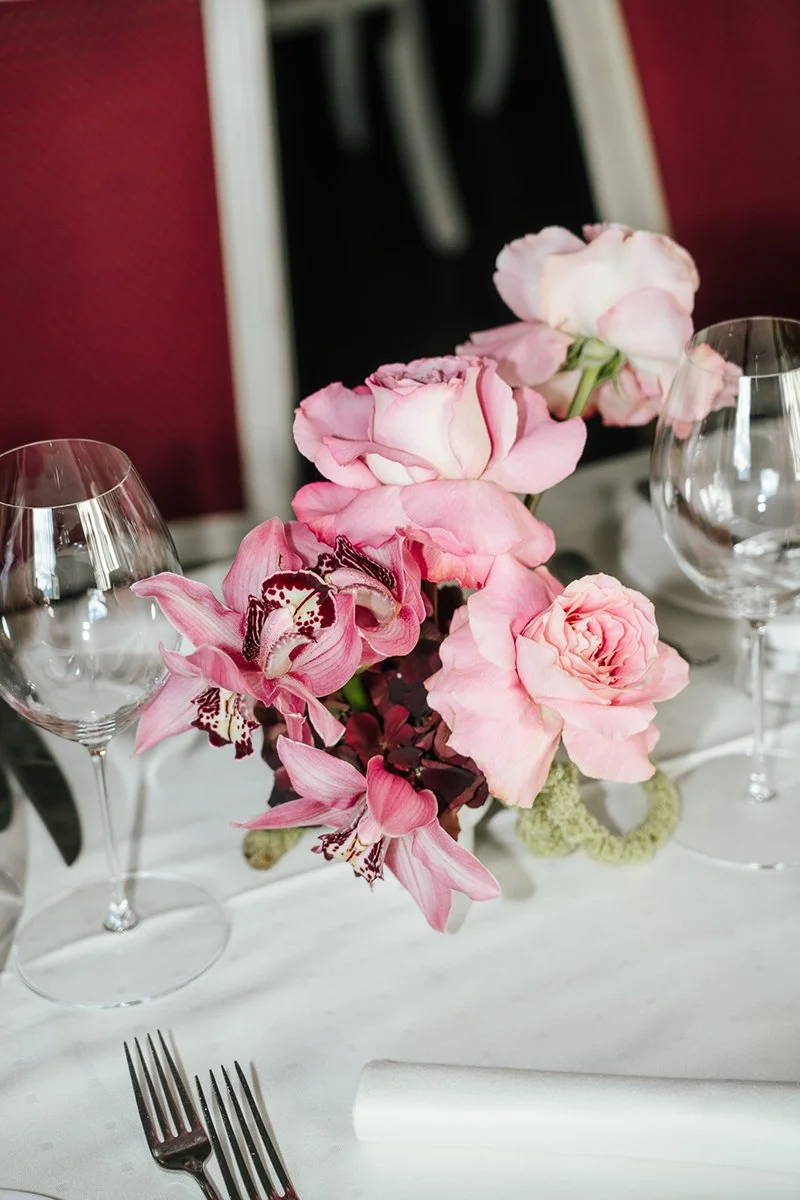 Ein pinkes Blumenarrangement steht auf einem weissen eingedeckten Tisch.
