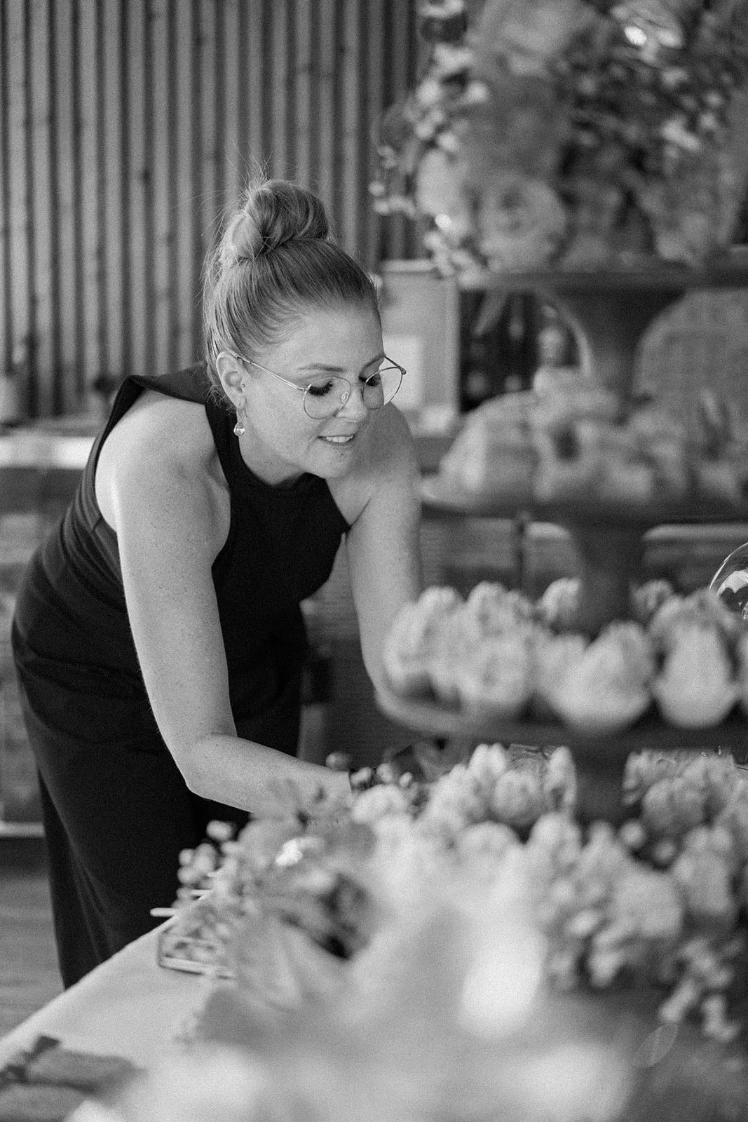 Frau in schwarzem Kleid und Brille arrangiert Blumen in einem Raum mit Holzwand