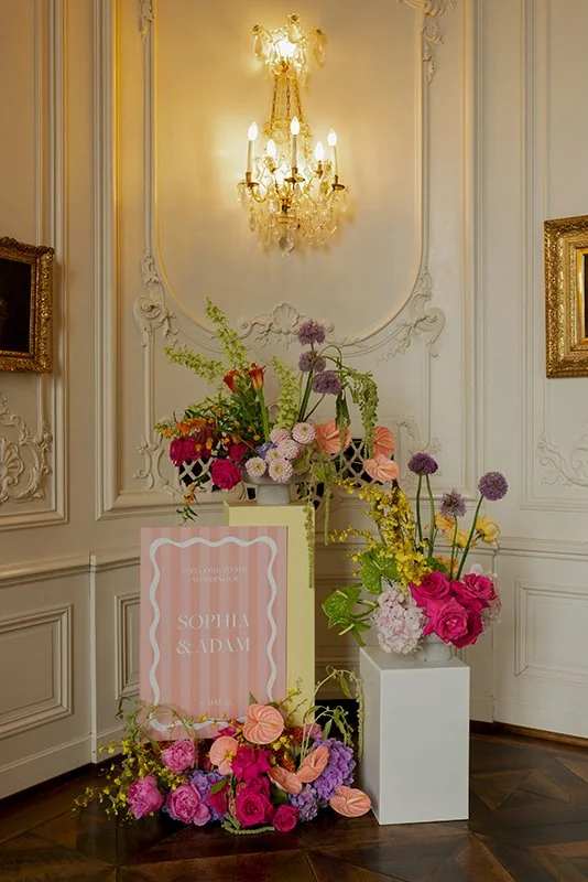 Verschiedene bunte Blumenarrangements sind auf Säulen angeordnet vor einer weissen Wand.