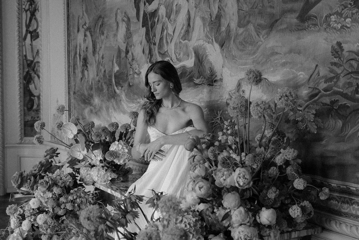 Eine Frau in einem weissen Kleid sitzt inmitten von vielen Blumen.