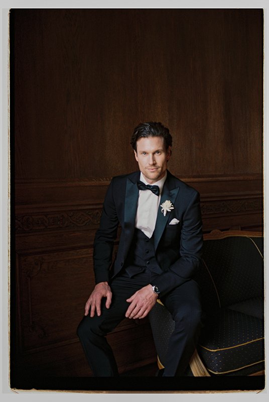 Ein Mann sitzt im schwarzen Anzug vor einer Holzwand.