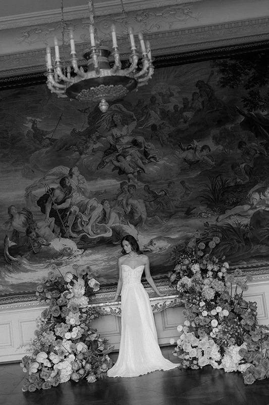 Eine braut steht vor einer Wand umgeben von bunten Blumen.