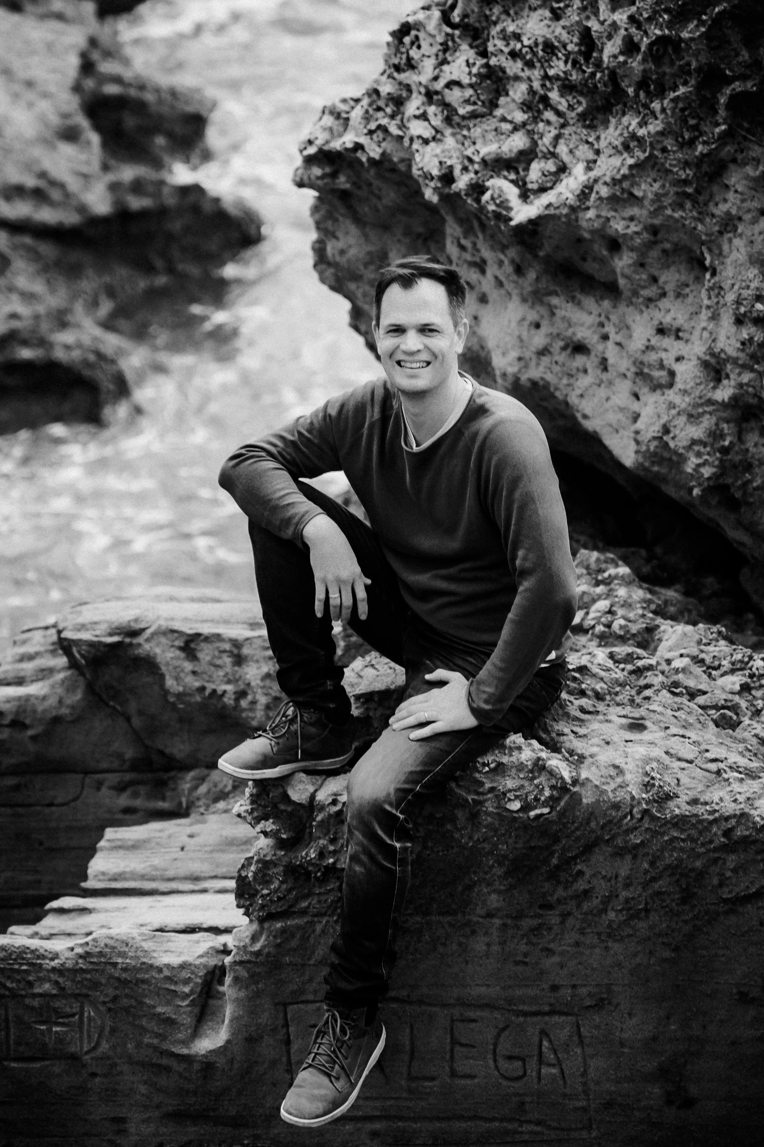 Ein lachender Mann sitzt auf einem Felsen am Meer, umgeben von Gestein und Wasser, in Schwarz-Weiß fotografiert.