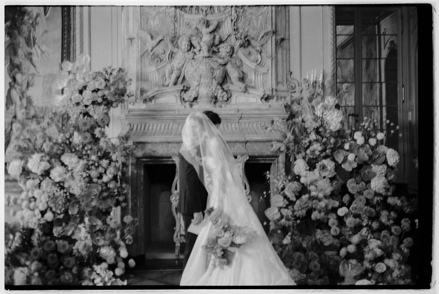 Ein Brautpaar steht vor einem Chemine umgeben von Blumen.